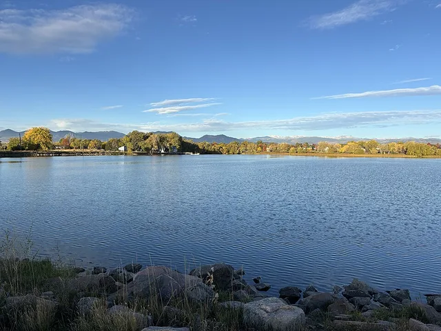 This photograph looks west from the east shore of Waneka Lake in Lafayette, Colorado, showing riprap in the foreground, calm water across the reservoir, and a line of cottonwoods...