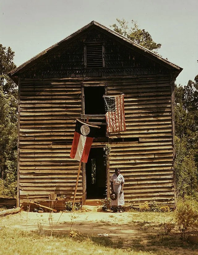 en:African American woman standing by building with en:Georgia state flag and en:United States flag near en:White Plains, Georgia , ca. 1941.