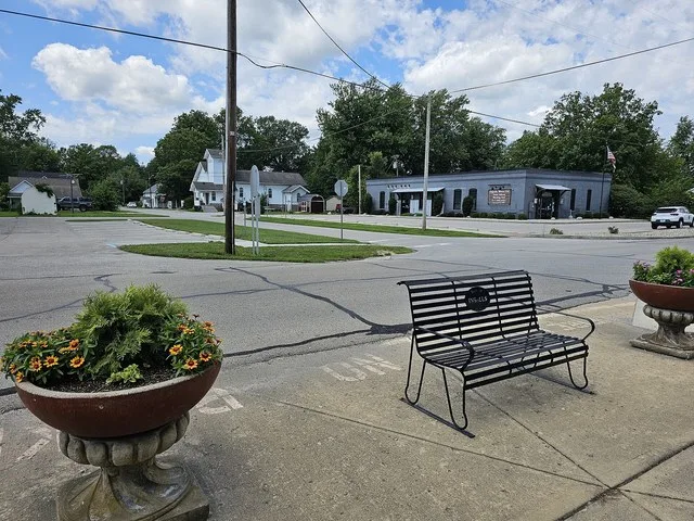 This image shows the town hall of Ingalls, Indiana. The foreground includes a bench with the word Ingalls on it, and some flower pots. Beyond the town hall is a white church.