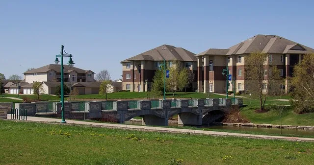A park trail and walking bridge by a pond near modern apartment buildings in the Liberty Centre area of North Liberty, Iowa, United States.