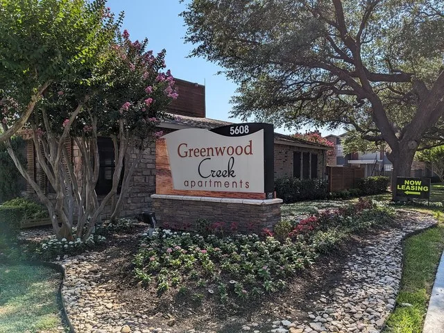 Greenwood Creek Apartments Entrance sign. It is within a flowerbed with summer flowers and river stones. Some crepe myrtles are visible as well as a building behind the sign that...