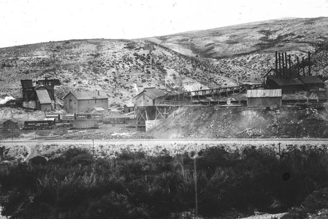 Exterior view of the Diamondville Coal Mine. Railroad tracks cross in front of the mine. There are smoke stacks connected to a tipple and other mine buildings on the right side. O...
